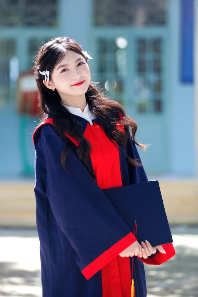 Smiling young woman in graduation robe holding diploma outdoors on a sunny day.
